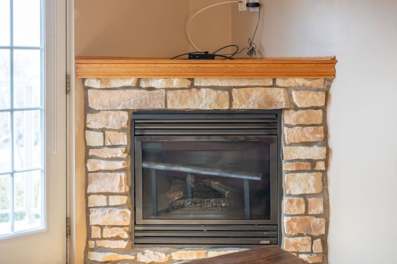 A stone fireplace with a wooden mantel, a power outlet above it, and a window to the left.