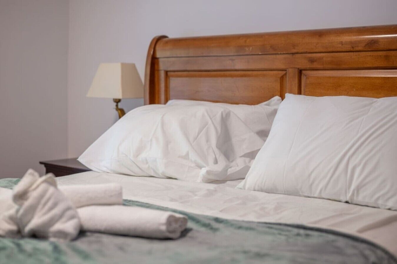 Ein ordentlich gemachtes Bett mit weißen Leinen und einem aufgerollten Handtuch, ein Holz-Kopfstück sowie ein Nachttisch mit Lampe.