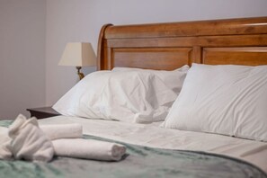 A neatly made bed with white linens and a rolled towel, a wooden headboard, and a bedside table with a lamp.