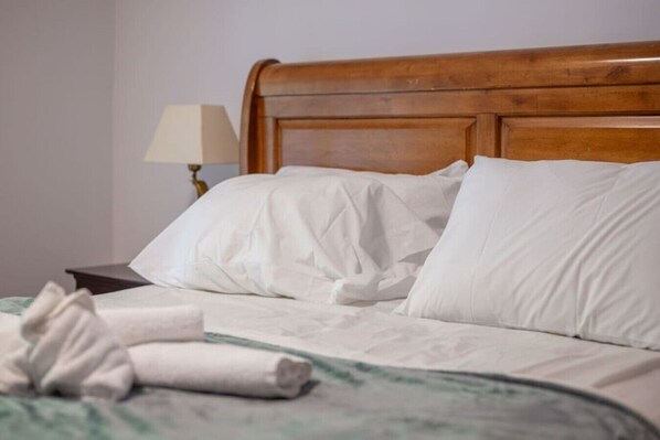 Una cama bien hecha con sábanas blancas y una toalla enrollada, un cabecero de madera y una mesita de noche con una lámpara.