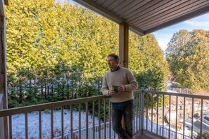 A person standing on a wooden deck with a railing, overlooking a snowy landscape and dense trees.