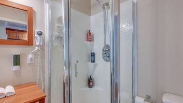 A glass-enclosed shower with a handheld showerhead, a handheld dryer, and a bottle of shampoo on a shelf.