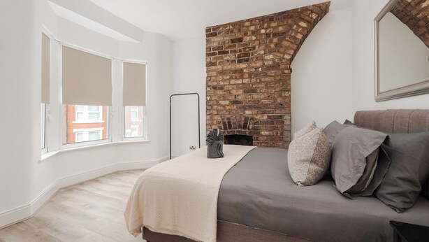 Een slaapkamer met een open haard van baksteen, een bed met grijze beddengoed, een raam met zonwering en een witte muur.