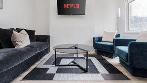 Een moderne woonkamer met een zwarte bank, een blauwe bank, een glazen salontafel, en een tv aan de muur waarop 'NETFLIX' staat.