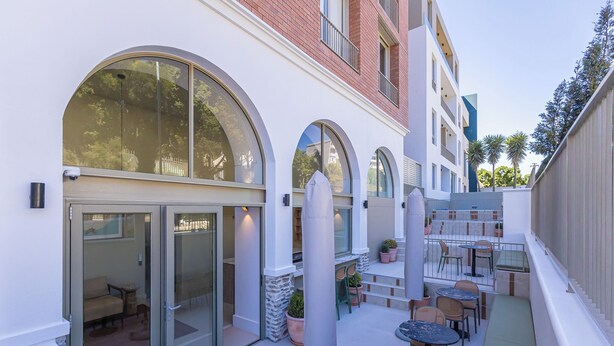 Un patio con mesas al aire libre, una entrada con puerta de vidrio corredera y fachada de edificio de ladrillo.