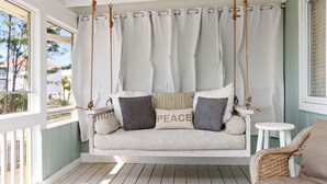 Une balançoire blanche, installée sur le porche et dotée d’un coussin sur lequel est inscrit le mot « PAIX », une chaise en osier et une vue sur l’extérieur.