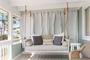 Eine weiße Verandaschaukel mit Sitzkissen, ein Korbstuhl und Blick ins Freie.