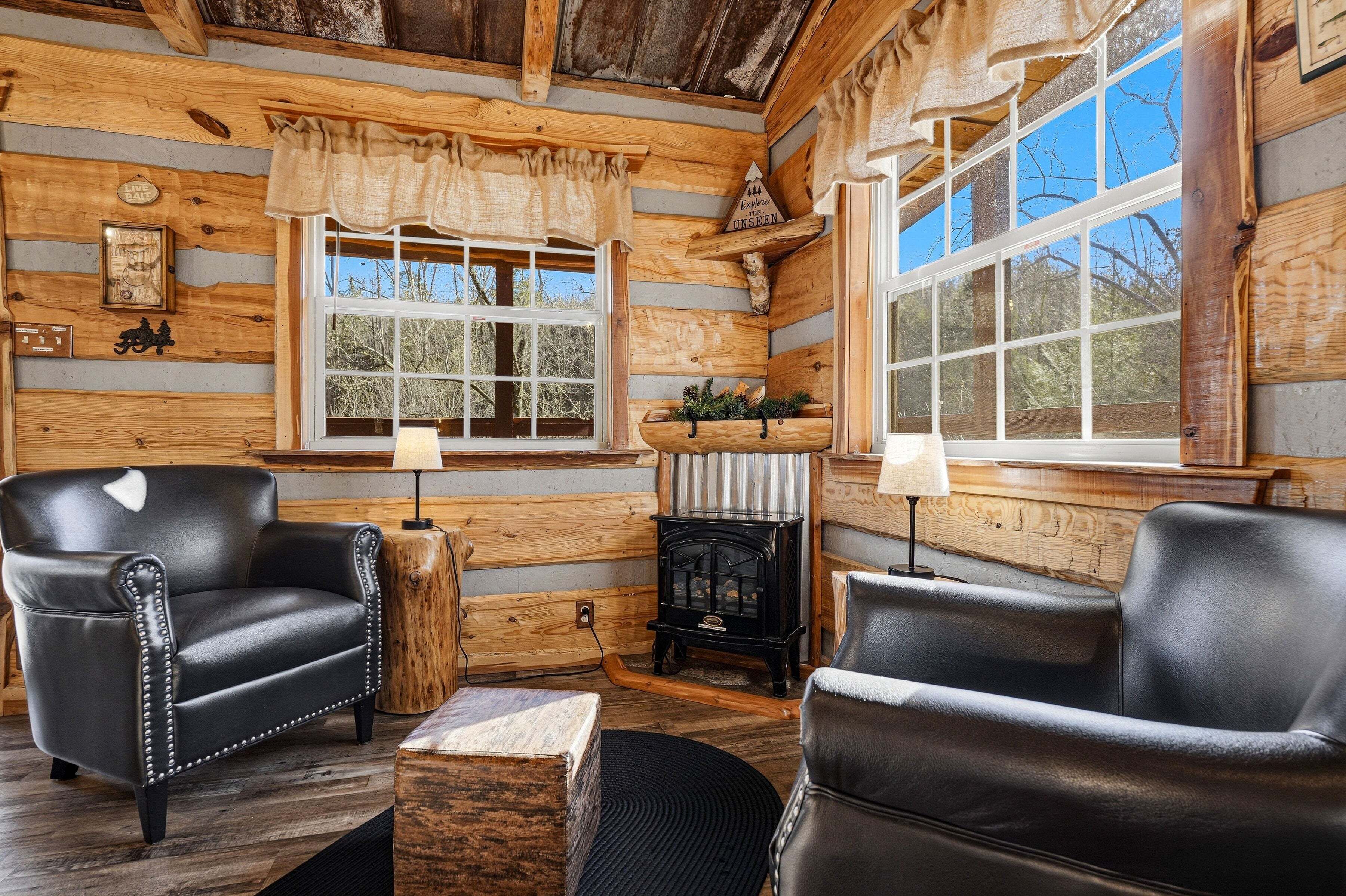 A log cabin interior with two leather armchairs, a wood-burning stove, and large windows.