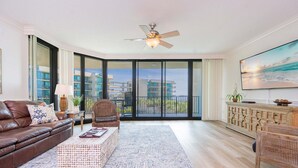A spacious living room with a large window, a ceiling fan, a flat-screen TV, and a wicker coffee table. - Phoenix On The Bay 1309 3 Bedroom Condo (Orange Beach)