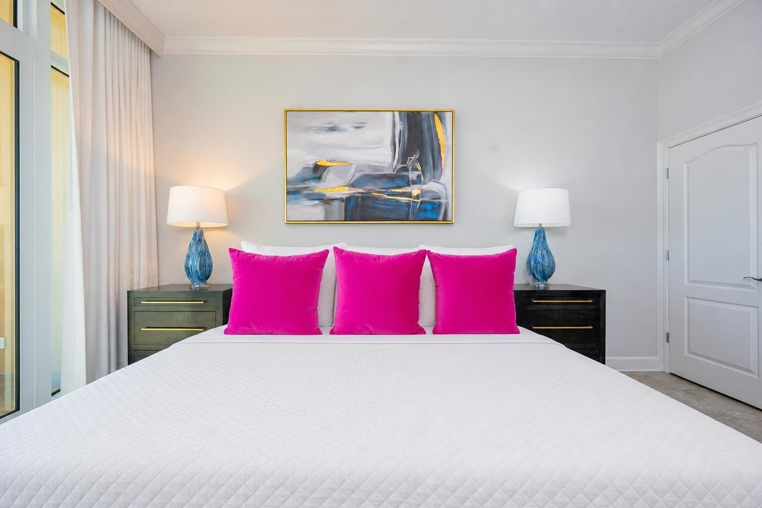 A hotel room with a large bed, two bedside tables with lamps, a painting on the wall, and a door to the right.