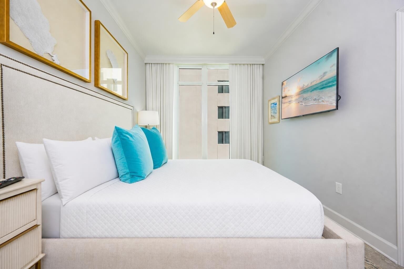 A hotel room with a bed, a nightstand, a ceiling fan, a painting, and a view of the outside.