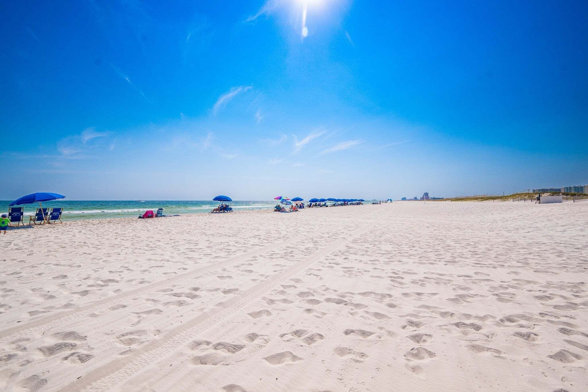 A wide sandy beach with footprints, a clear blue sky, and distant buildings.