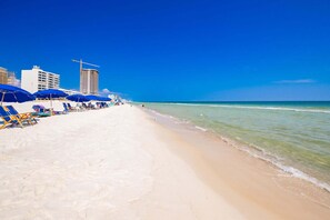 Een strand met ligbedden en parasols, een heldere blauwe lucht en gebouwen op de achtergrond.