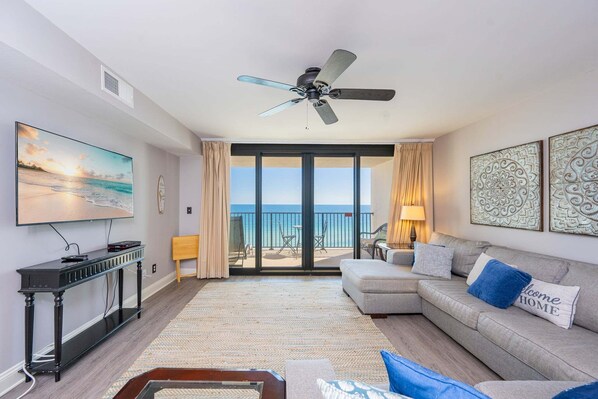 A living room with a sofa, a TV, a ceiling fan, and a balcony with a view of the ocean. - Wind Drift 604s 2 Bedroom Condo (Orange Beach)