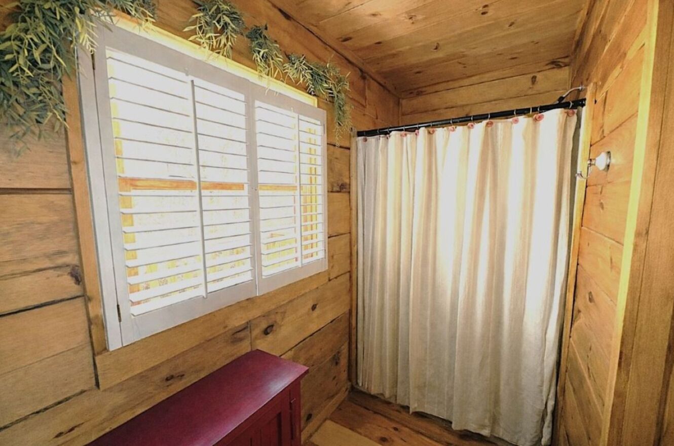 Una cabaña de madera con ventana, persianas, una cortina y un pequeño banco rojo.