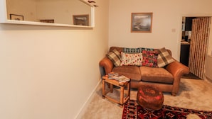 A living room with a brown sofa, a small wooden table, a round ottoman, a patterned rug, a mirror with antlers, and a framed picture on the wall. - 25 Low Seaton (Workington)