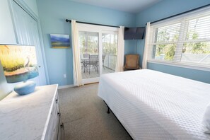 Un dormitorio con cama, escritorio, silla, televisor y puerta corrediza de cristal a un balcón.