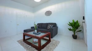 A living room with a dark sofa, a glass coffee table, a potted plant, and a decorative wall piece.