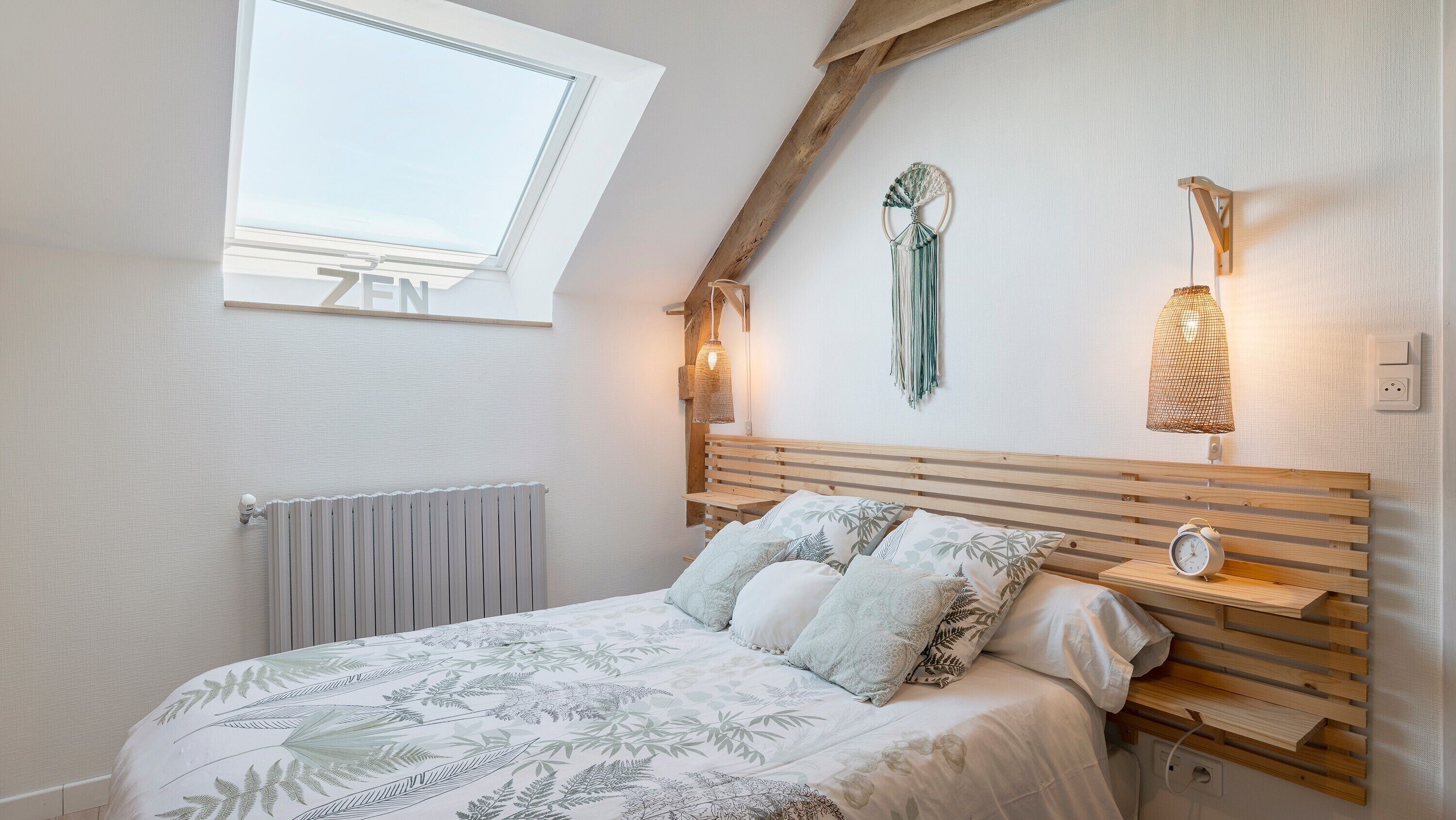 Une chambre avec un plafond en pente, un puits de lumière, un lit avec une literie à motifs, une table de chevet avec une horloge et des lampes fixées au mur.