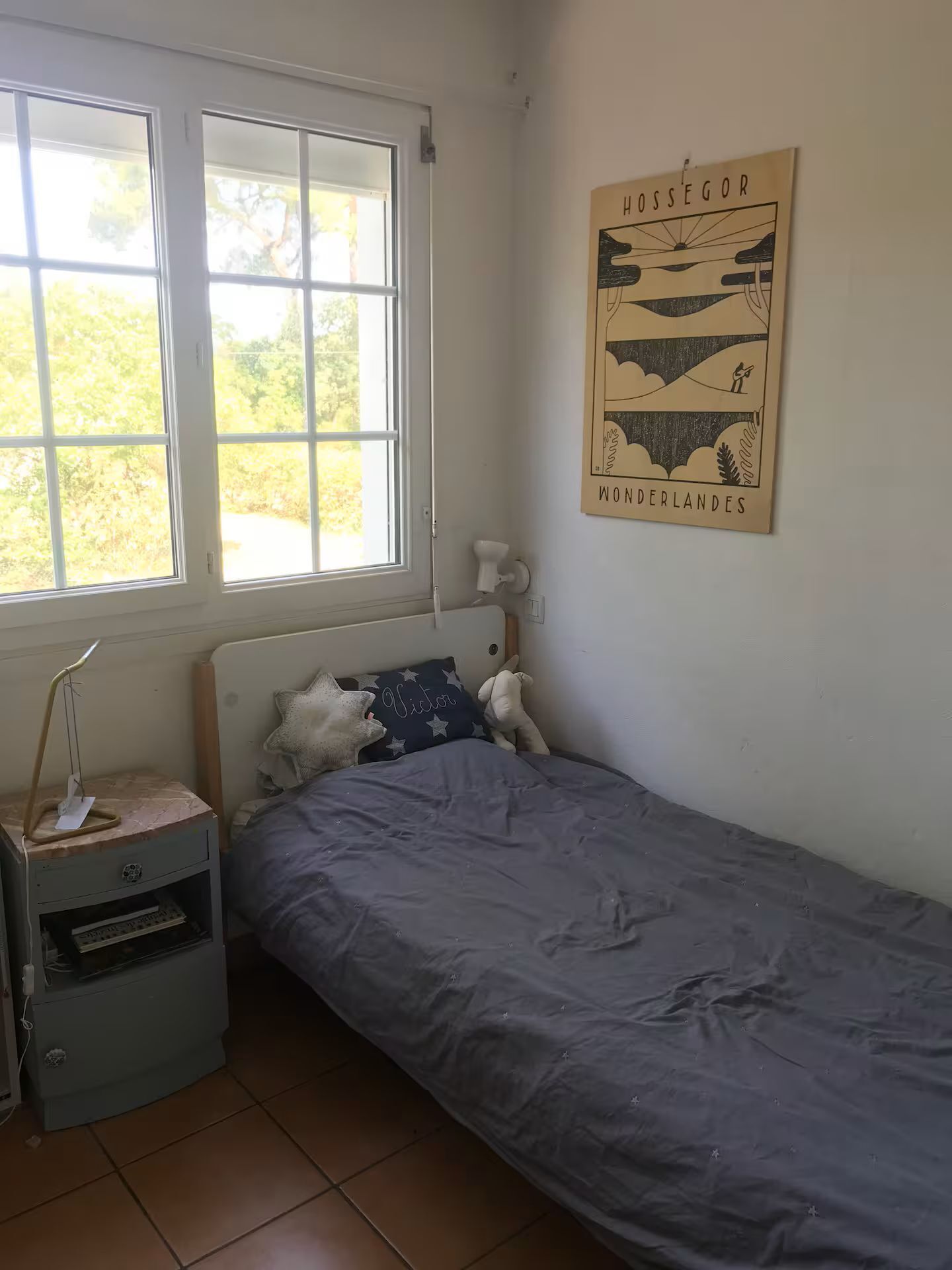 A small, single-bed room with a window, a bedside lamp, a wall-mounted lamp, and a poster.