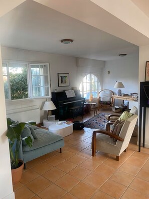 Ein Wohnzimmer mit einem Klavier, einem Sofa, einem Sessel, einer Lampe und einem großen Fenster.