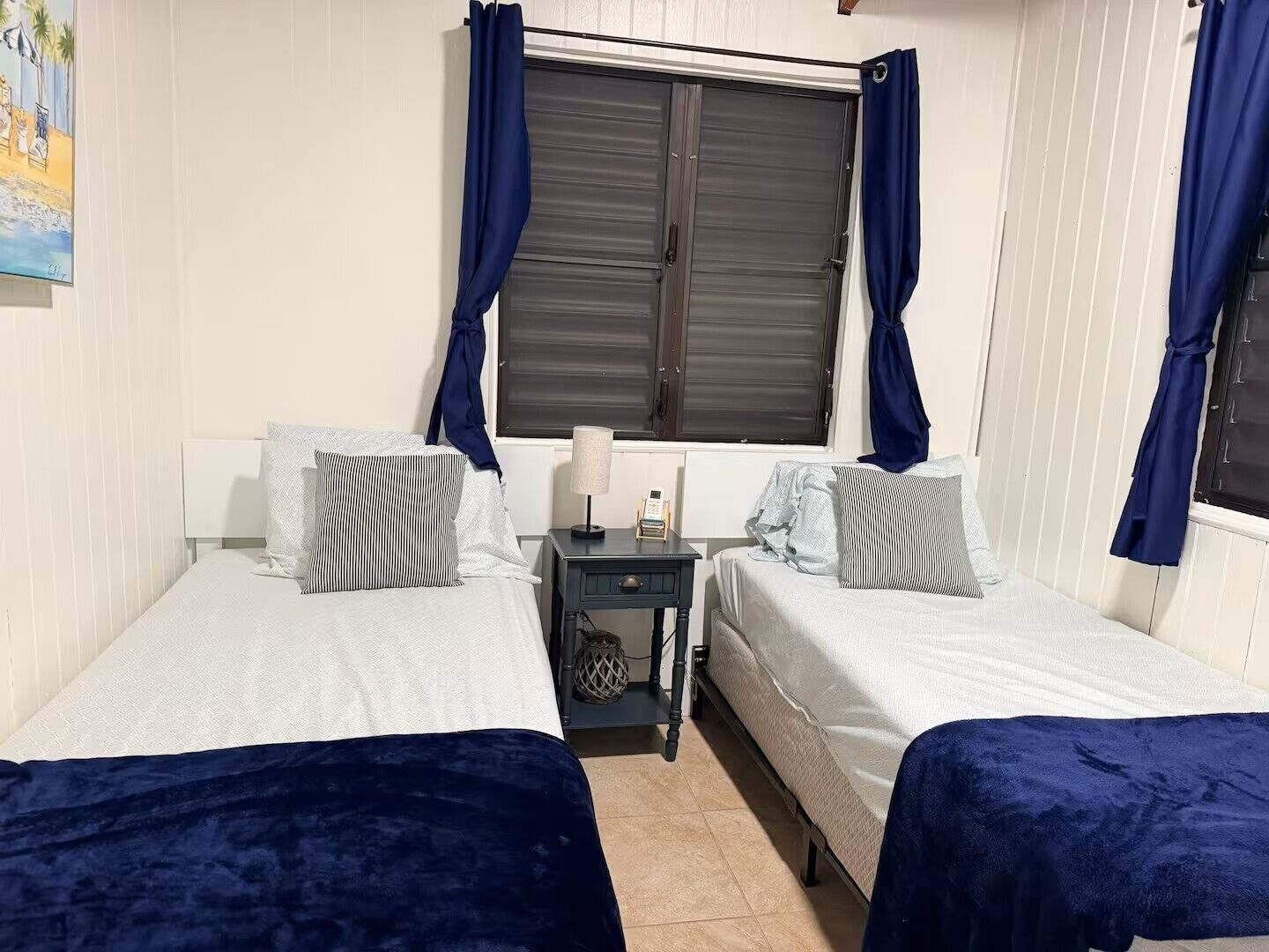 A room with two beds, a nightstand, a lamp, and a window with curtains.