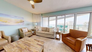 A spacious living room with a large window, a ceiling fan, and a painting on the wall. - Tides 1311 - Gulf Views | Beachfront Resort Pools (Miramar Beach)