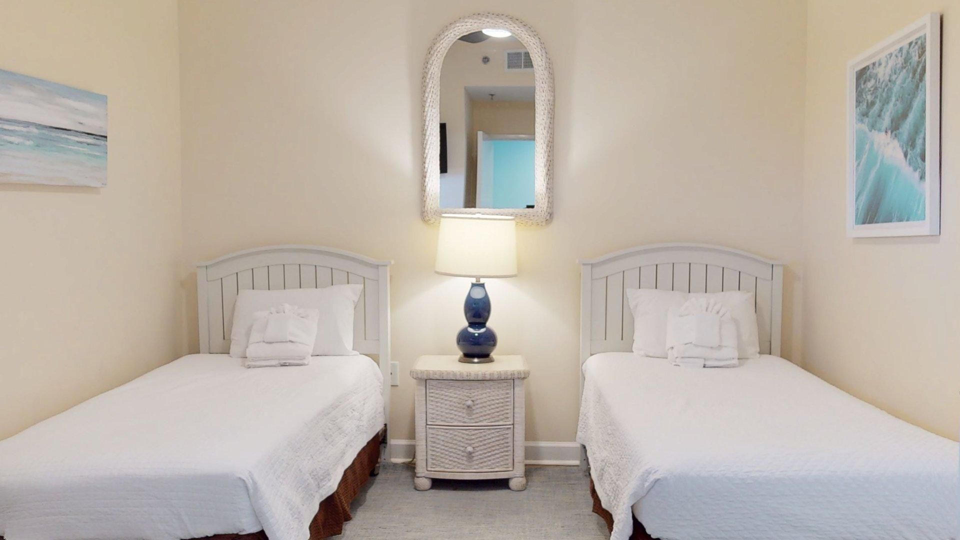 Quarto de hotel com duas camas de solteiro, mesa de cabeceira com abajur, espelho e obras de arte nas paredes.