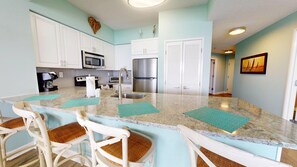 A kitchen with white cabinets, a stainless steel refrigerator, a microwave, and a coffee maker. There are two white chairs with woven seats. - Tides 1311 - Gulf Views | Beachfront Resort Pools (Miramar Beach)