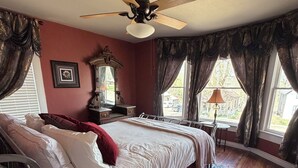 Une chambre à coucher avec un lit, un ventilateur de plafond, une fenêtre avec des rideaux, une table de chevet et une lampe.