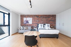 A modern bedroom with a brick wall, a bed, a sofa, a dining table, and a large window. - Sleek Sanctuary in the Sky (Cape Town)