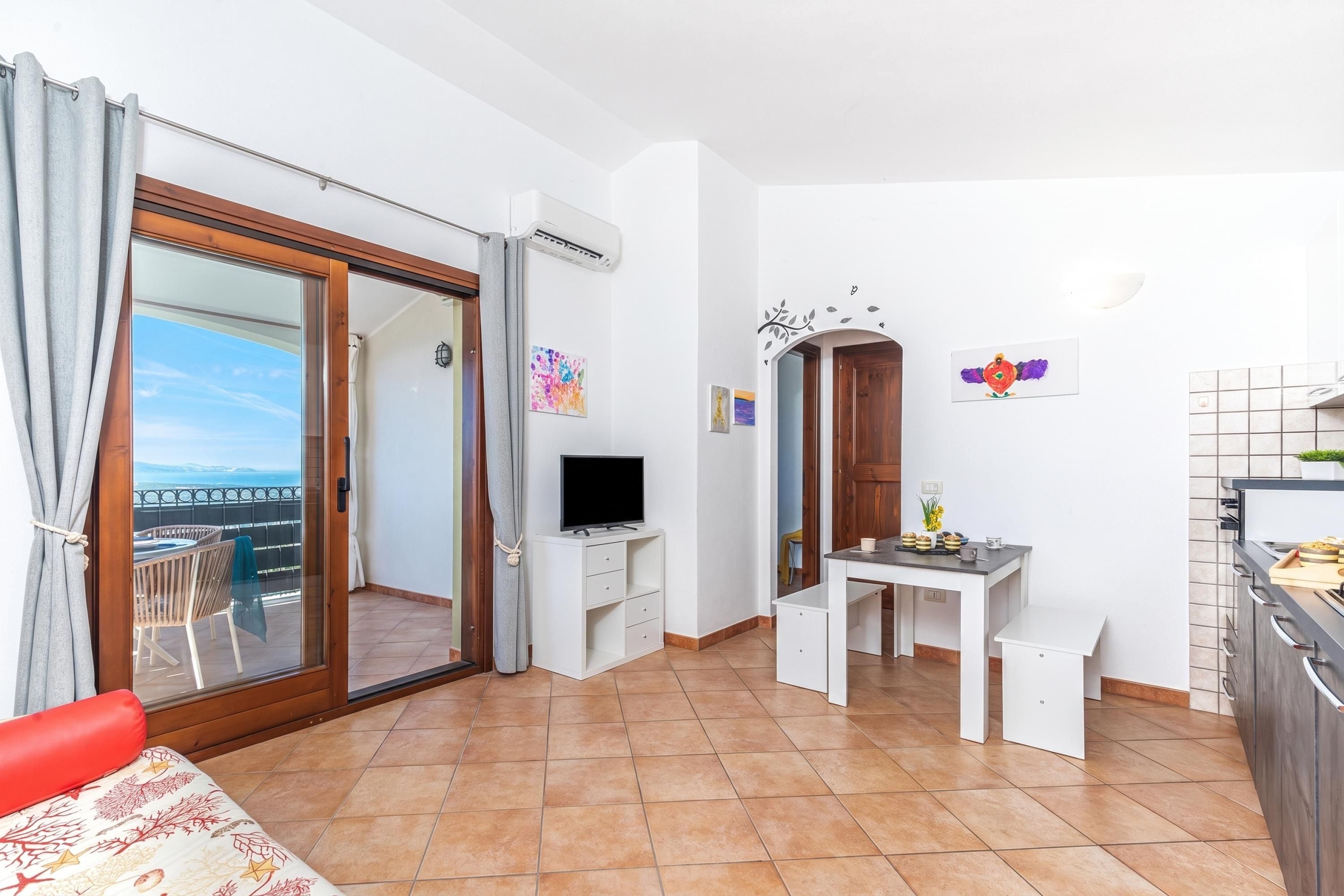 Una stanza con una porta vetrata scorrevole che dà su un balcone, una piccola scrivania bianca con una TV a schermo piatto, un tavolo da pranzo con sedie e un angolo cottura con rivestimento piastrellato.