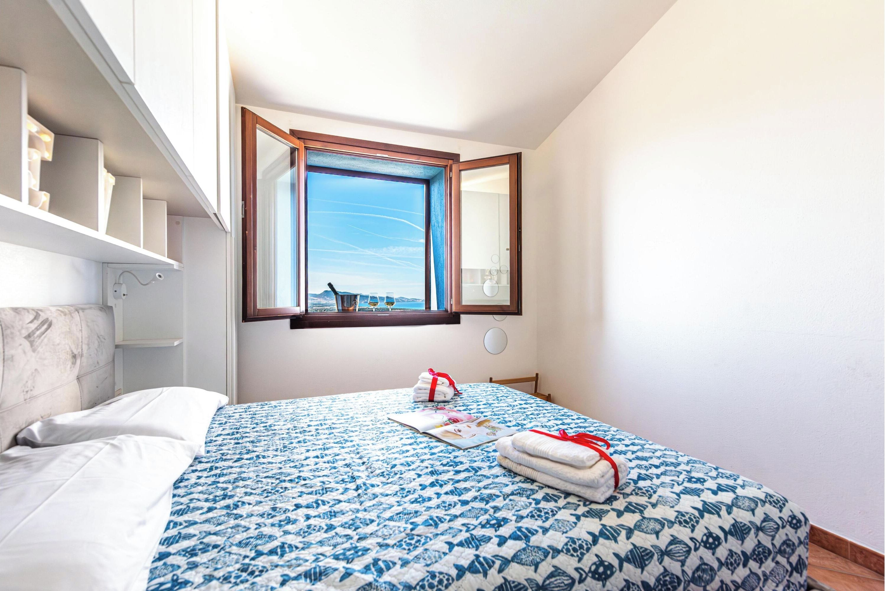 A bedroom with a bed, a window with a view, a bedside table, and a wall-mounted shelf.