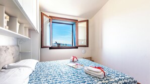 A bedroom with a bed, a window with a view, a bedside table, and a wall-mounted shelf. - Vhouse (Badesi)
