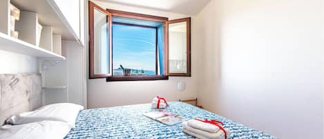 A bedroom with a bed, a window with a view, a bedside table, and a wall-mounted shelf.