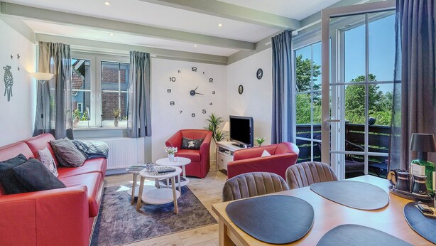 Ein modernes Wohnzimmer mit einer Couch, Sesseln, einem Couchtisch, einem Fernseher und einem großen Fenster mit Vorhängen.