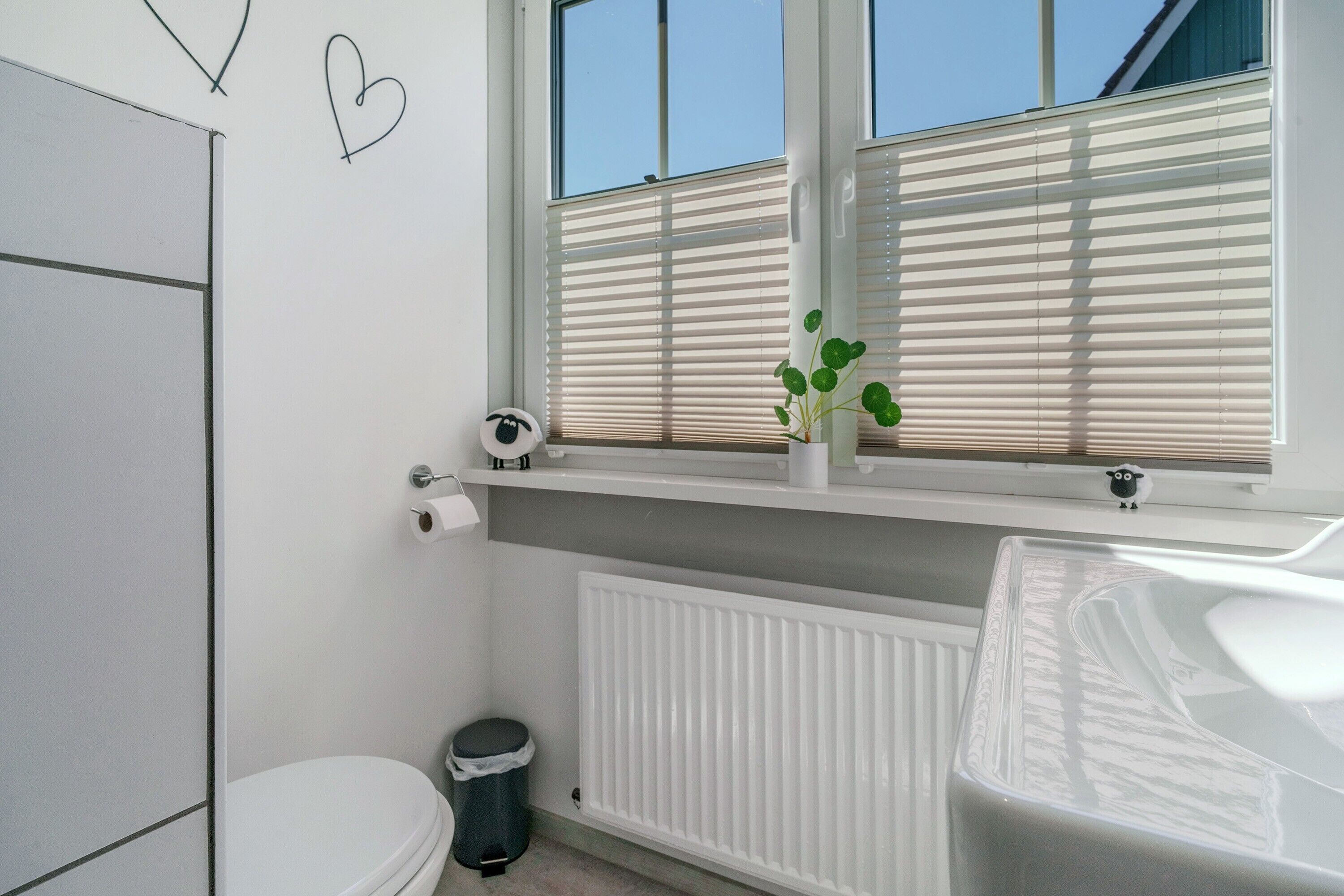 Ein Badezimmer mit Toilette, Heizkörper, Fenster mit Jalousien und einer kleinen Ablage mit einer Pflanze.