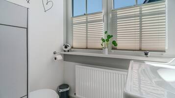 Ein Badezimmer mit Toilette, Heizkörper, Fenster mit Jalousien und einer kleinen Ablage mit einer Pflanze.