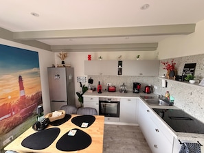 A modern kitchen with a refrigerator, stove, and sink. There is a dining table with black placemats and a painting of a lighthouse.