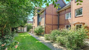 Un immeuble résidentiel à la façade en briques, doté d’un chemin pavé et d’un jardin bien entretenu où poussent diverses plantes et fleurs.