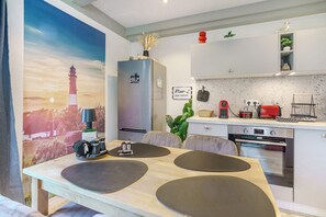 A modern kitchen with a built-in oven, a dining area with a table and chairs, and a large painting of a lighthouse. - Apartment Schwarzes Schaf – Central Location, Cozy Ambience, Big Heart (Pellworm)