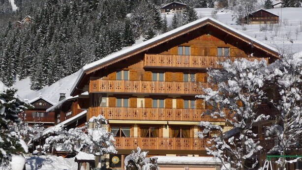 Un edificio de madera grande con varios balcones, rodeado de árboles cubiertos de nieve y un telón de fondo montañoso.