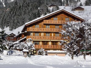 A large wooden building with multiple balconies, surrounded by snow-covered trees and a mountainous backdrop. - Location Appartement 8 personnes au center de LA CHAPELLE D'ABONDANCE (parking) (La Chapelle-d'Abondance)