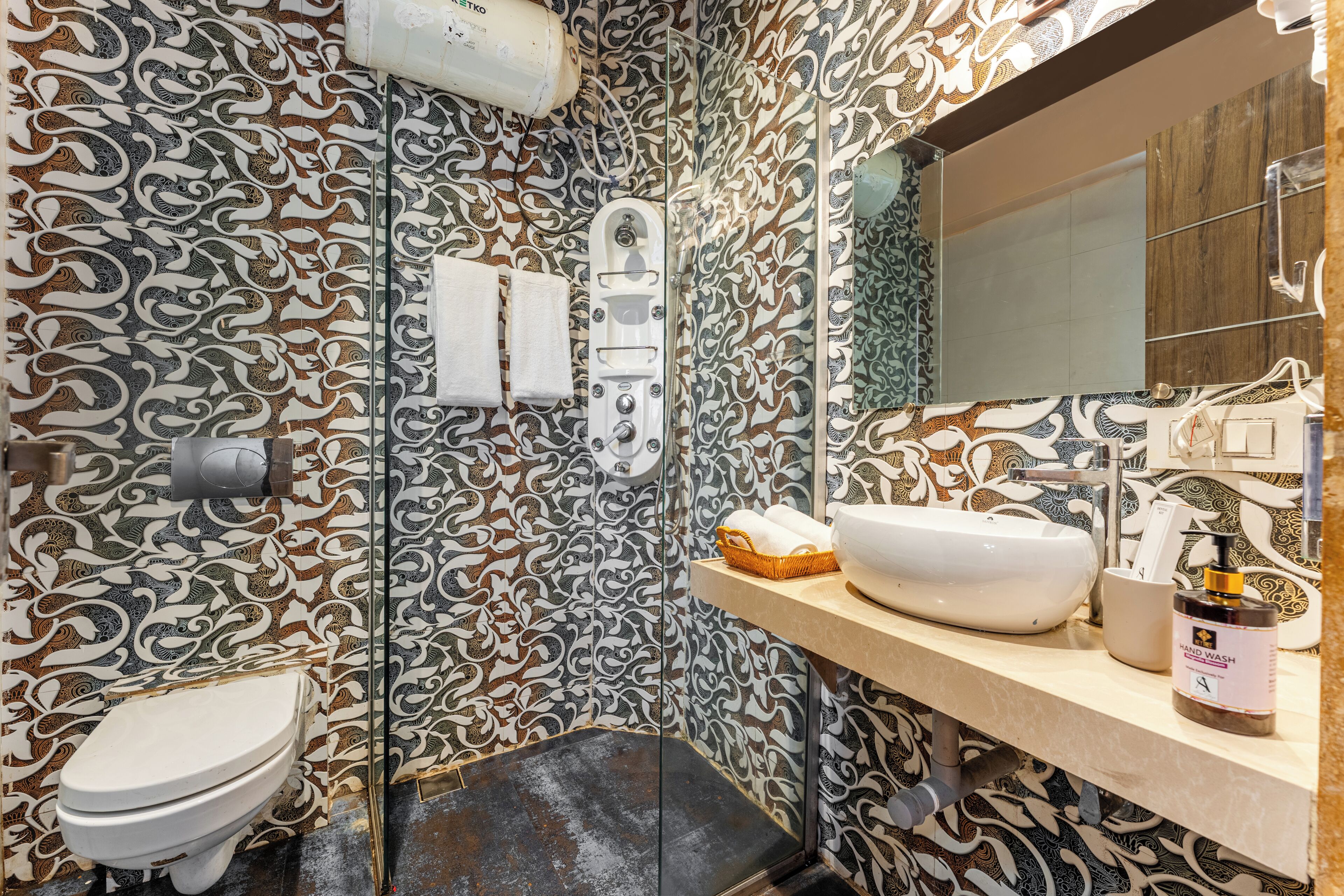 Une salle de bain avec des carreaux décoratifs, des toilettes, un bidet, un lavabo avec un flacon de savon liquide et un miroir.