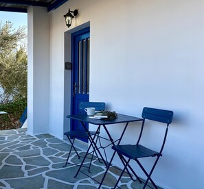 A blue door with a keyhole, a small table with a cup and a book, and two blue chairs on a stone-paved porch. - Origins II | Calm Studio near Beach & Taverns (Piso Livadi Paros)