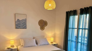 Ein ordentlich bezogenes Bett mit Kissen, zwei Nachttische mit Lampen, ein an der Wand hängendes Kunstwerk, eine Wanddekoration aus Gewebe und ein Fenster mit Vorhängen.
