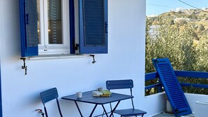 Ein Balkon mit Tisch und Stühlen, ein Fenster mit blauen Fensterläden und Blick auf eine Hügelkuppe.
