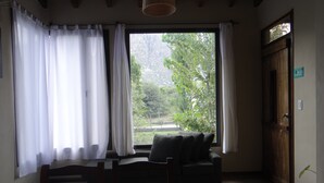 Une pièce avec un lit, une chaise, une fenêtre avec des rideaux et une vue sur la verdure à l’extérieur.