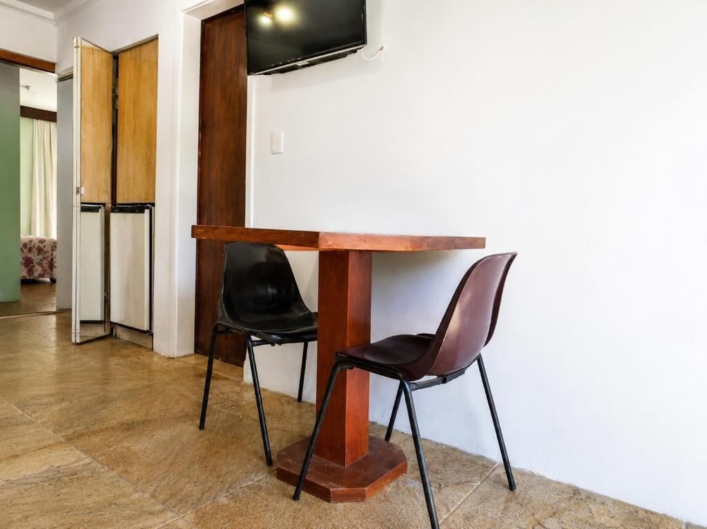 Een kamer met een houten tafel en twee stoelen, een aan de muur bevestigde televisie, en een deur die naar een andere kamer leidt.