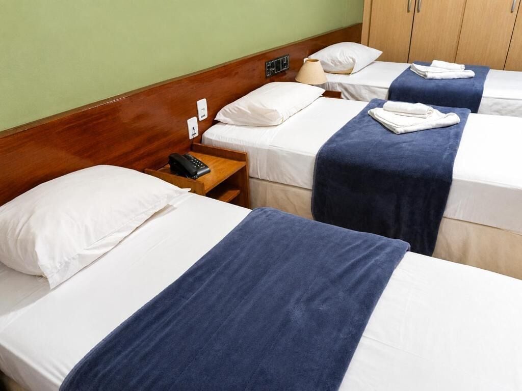 Habitación de hotel con tres camas individuales, cada una con ropa blanca y una manta azul. Cuenta con un cabecero de madera, una mesita de noche con lámpara y un teléfono.